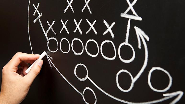 Hand diagramming football play in chalk on chalkboard.